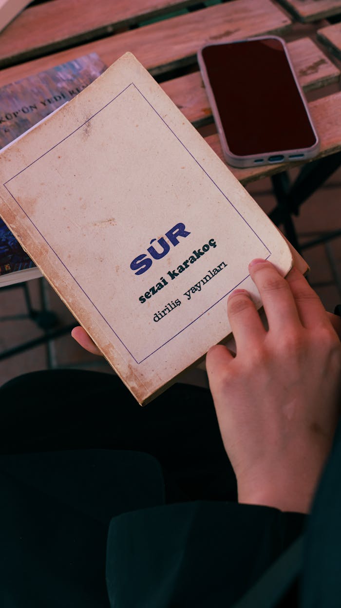 Close-up of a person reading the book Sür by Sezai Karakoç in an outdoor café.